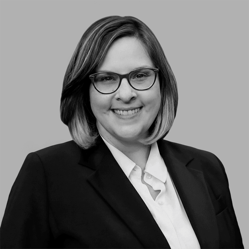 Smiling person with glasses and short hair, wearing a suit jacket and white shirt, on a plain background.
