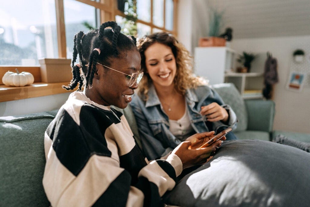 Two friends sitting on a couch, smiling and looking at a smartphone together in a bright living room.