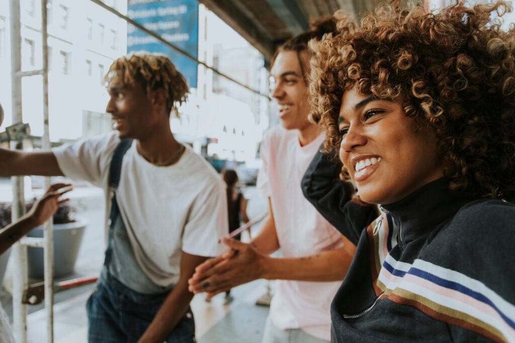 Three young adults smiling and talking outdoors on a city street, with one person in the foreground wearing a striped jacket.